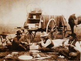 A black and white historical photo of cowhands eating on the ground in camp in front of a chuckwagon